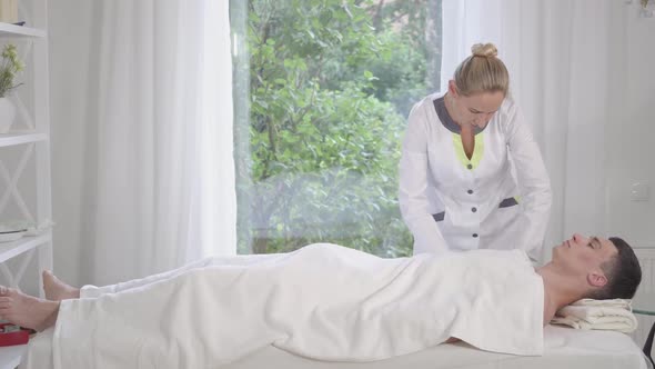 Serious Professional Doctor Massaging Body of Young Brunette Man Covered with White Towel. Side View