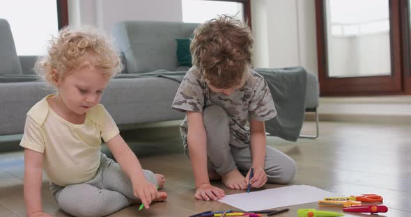 A Little Boy and a Little Girl are Drawing with Pencils on on the White Paper on the Floor Looking