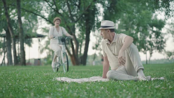 Handsome Mid-adult Caucasian Man Sitting on Green Lawn in Summer Park and Waving To Cheerful Woman