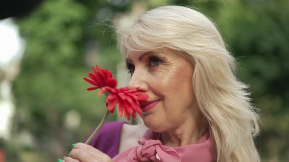 Closeup Blond Adult Woman Smelling Red Flower Smiling Looking Up Standing in Summer Park Outdoors