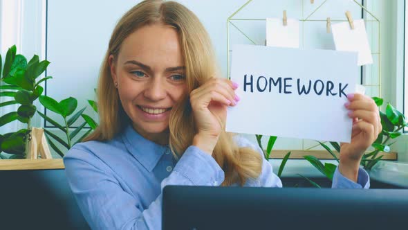 Young Female Teacher Sitting at Desk and Using Laptop Showing HOMEWORK Text at Home