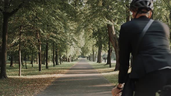 Young Man Riding a Bike in City Park Wearing Business Suit and Helmet Back View