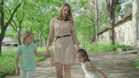 Front View of Young Charming Mother Holding Hands of Cheerful Daughter and Son and Walking Along the