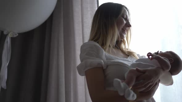 Portrait of Loving Caucasian Woman with Newborn Baby in Hands Standing at Window Indoors Admiring