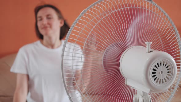 Young Woman Sits in Front of Electric Fan Waving Hands Air Stream Blowing Hair