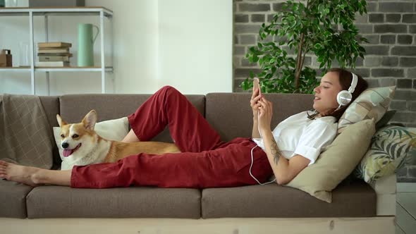 Young Young Woman Listening to Music and Using Gadget on Couch with Dog in Living Room Avki