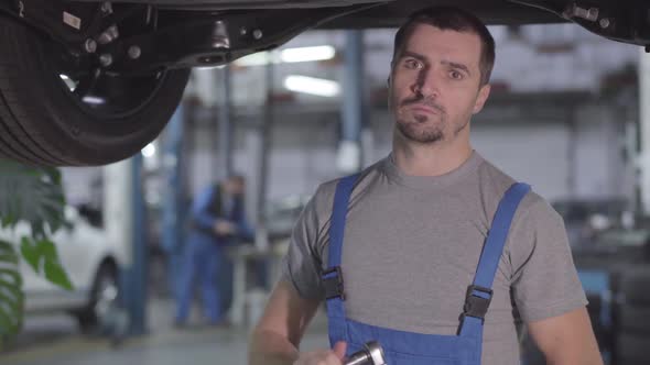 Portrait of Young Serious Caucasian Man in Blue Workrobe Standing Under Car in Repair Shop and