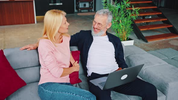 Lovely Middle Aged Couple Man and Woman Talking While Sitting on Sofa in Living Room Holding Laptop