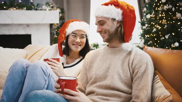 Mixed Race Pair Smiling and Cuddling Holding Red Mugs Sitting on Sofa at Home Against Decorated