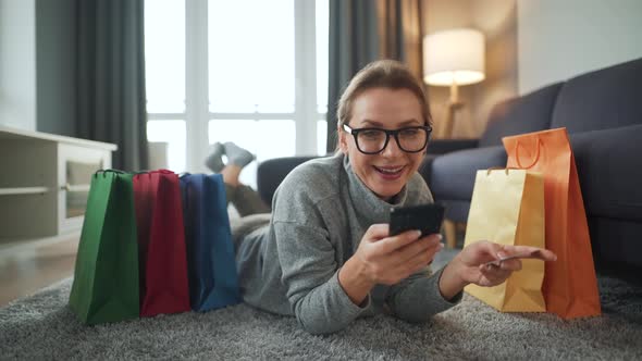 Happy Woman is Lying on the Floor and Makes an Online Purchase Using a Credit Card and Smartphone