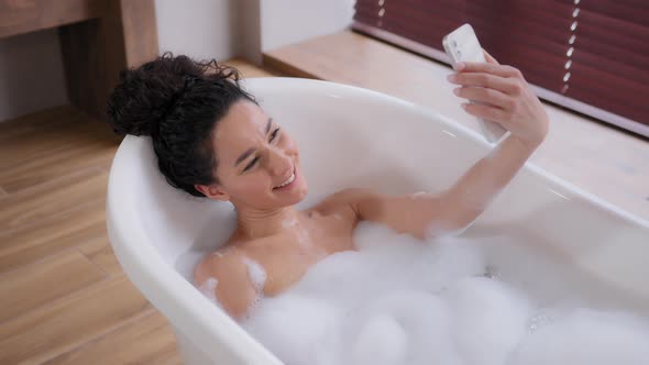 Young Happy Hispanic Woman Blogger Relaxing in Luxurious Bubble Bath Photographed on Phone Taking