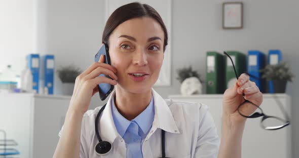 Physician in White Coat with Stethoscope Sitting at Desk and Talking on Smartphone