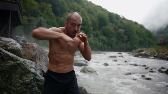 an Unshaven Man with a Bare Torso Trains By the River Against the Background of Mountains