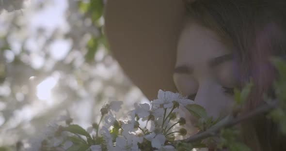 Closeup of Girl Wearing a Hat and Smelling the Flowering Branch of Apple Tree