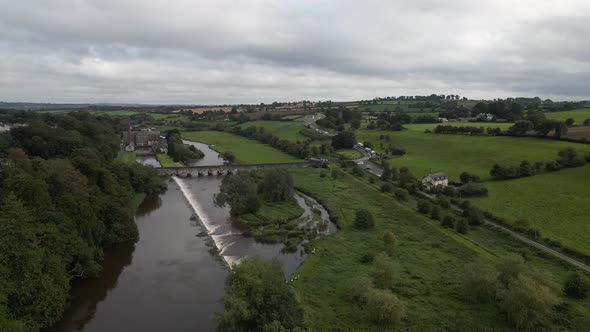 Drone shot of a large river flowing underneath an old bridge surrounded by green fields and trees.