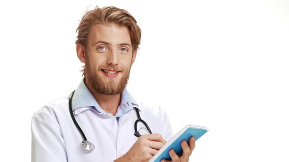 Glad Caucasian Male Doctor with Light Beard Wearing Medical Overall Smiling Making Notes in Blue