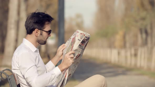 Man Sitting On Bench And Reading Newspaper. Handsome Guy Sits On Bench In Park And Reads Newspaper.