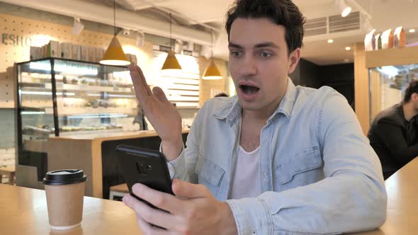 Shocked Young Man Wondering While Using Smartphone in Cafe
