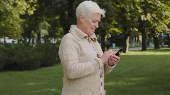 Grayhaired Retired Woman Walking in Park in Cool Weather Outdoor Using Phone Browsing Smartphone