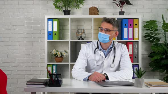 Doctor wears protective mask on face, white coat invites patient to sit on chair