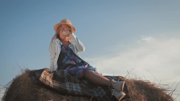 Pretty Girl in Blue Dress Drinking Milk on a Haystack in the Field. Child on a Summer Picnic.