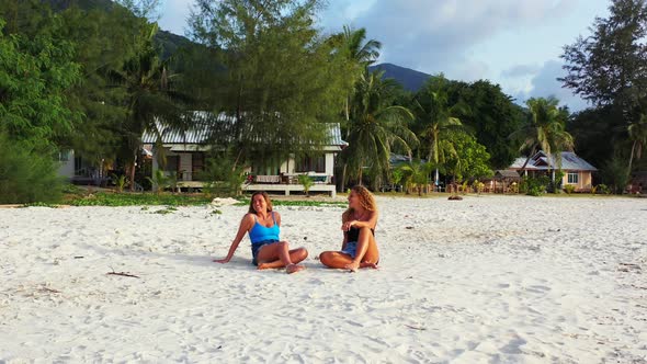 Smiling girls laughing and talking, sitting on white sandy beach of tropical island with palm trees