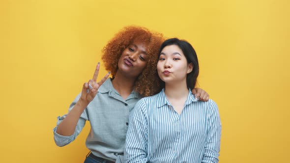 African American Black Woman and Her Asian Friend Posing Over the Yellow Background with Peace