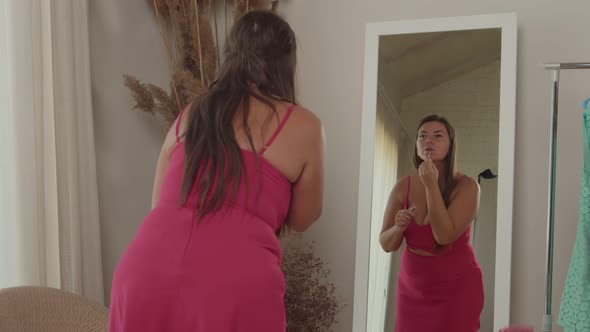 Plump Woman in Pink Dress Putting on Lipstick in Front Mirror Indoors