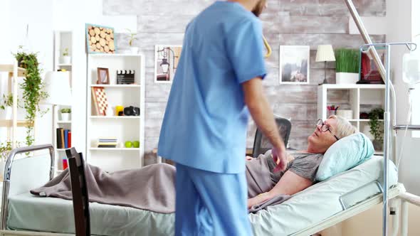 Male Assistant Taking a Seat Near Hospital Bed of Ill Lady Lying in Nursing Home