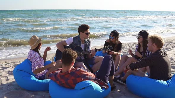 Young Man Playing Guitar Among Group of Friends Sitting on Easychairs on the Beach and Singing on a