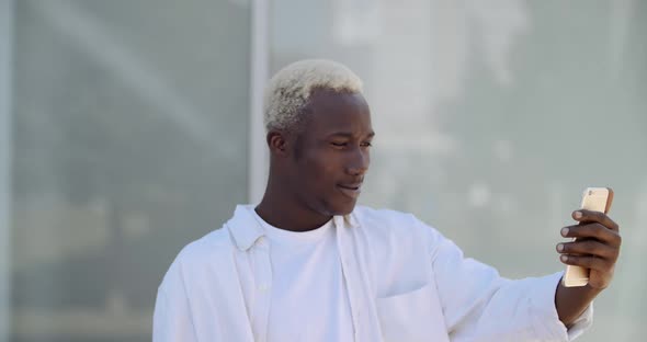 Young African American Hipster Guy in White Clothes Stands on Street Outdoors Alone, Holds Mobile