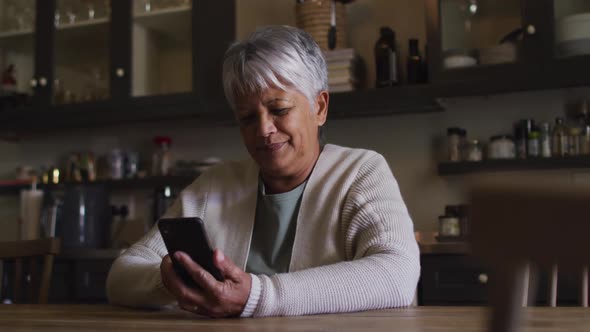 Smiling senior mixed race woman using smartphone sitting in kitchen