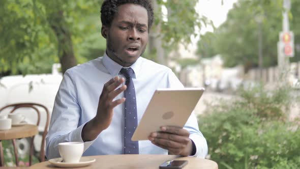 African Businessman Upset By Loss on Tablet Sitting in Outdoor Cafe