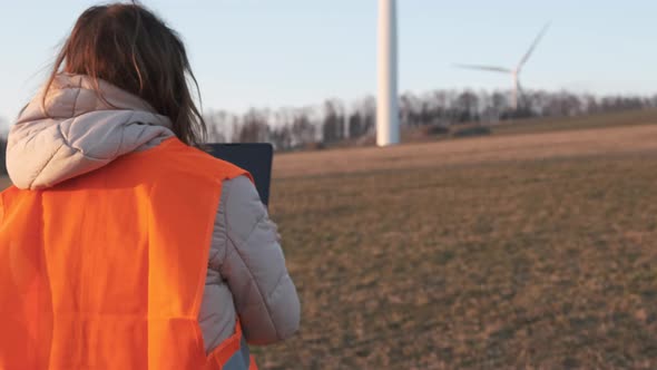 Female Engineer in Orange Vesta Goes to Wind Turbines with a Tablet to Checks Their Operation