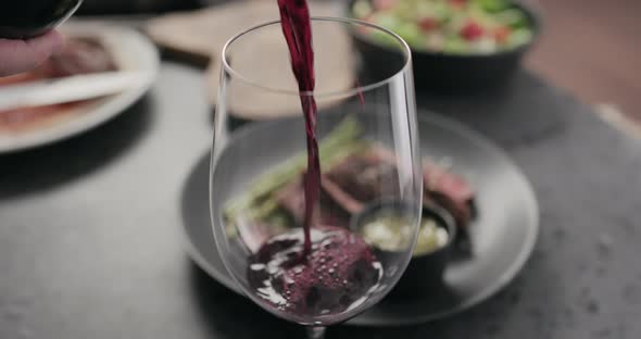 Slow Motion Closeup Pour Red Wine in Wineglass with Steak on Background