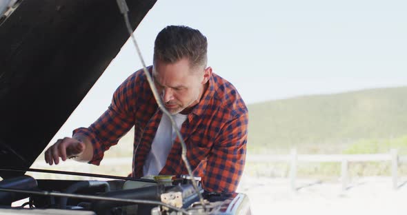 Stressed caucasian man looking at broken down car with open bonnet on sunny day at the beach