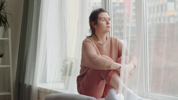 Contemplating Woman Sitting on Sill at Home and Looking through Window