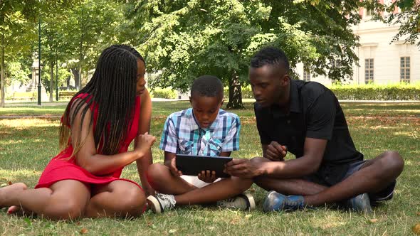 A Young Black Family Sits on Grass in a Park and Works on a Tablet