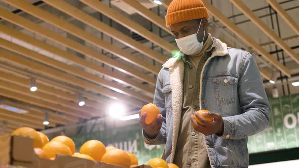 AfricanAmerican Male Shopper Wearing a Medical Mask Picks Out Fresh Oranges and Puts Them in a Bag