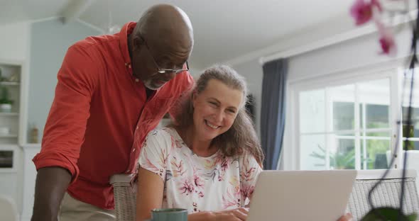 Happy senior diverse couple wearing shirts and using laptop in living room