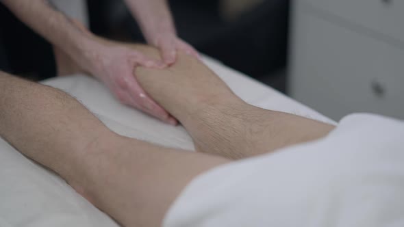 Hands of Professional Male Masseur Massaging Legs of Unrecognizable Man Lying on Massage Bed Indoors