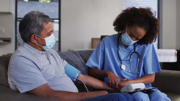 Mixed race female doctor wearing mask and senior man taking blood pressure at home