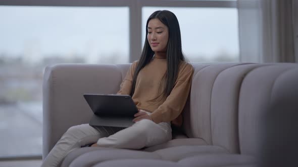 Wide Shot of Confident Charming Asian Woman Surfing Internet on Tablet Sitting on Couch in Home