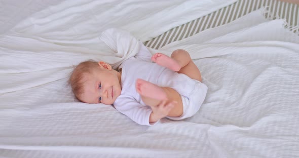 Pretty Baby is Playing with Arms and Legs Lying in a Bodysuit on a White Bedspread