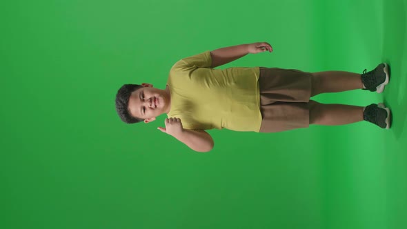 Full Body Of Happy Asian Little Boy Showing Thumbs Up Gesture While Standing On Green Screen