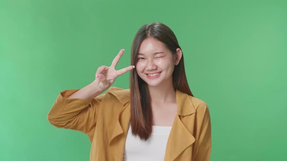 A Smiling Young Asian Woman Showing Gesture Peace While Standing On Green Screen In The Studio