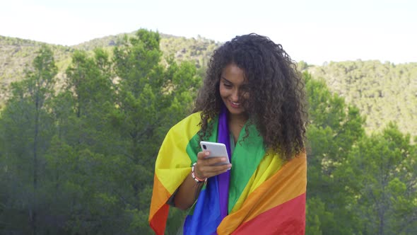 Curly haired woman typing with one hand on the phone and smiling while carrying an LGBT flag