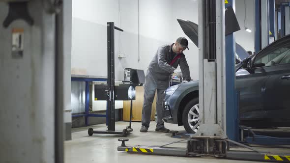 Adult Professional Auto Mechanic Adjusting Lightning Equipment in Auto Repair Shop. Caucasian Man