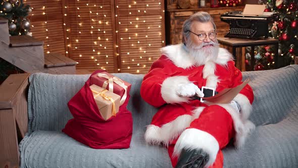 Smiling Male Santa Claus Blogger Reading Christmas Gift Wish List at Xmas Tree