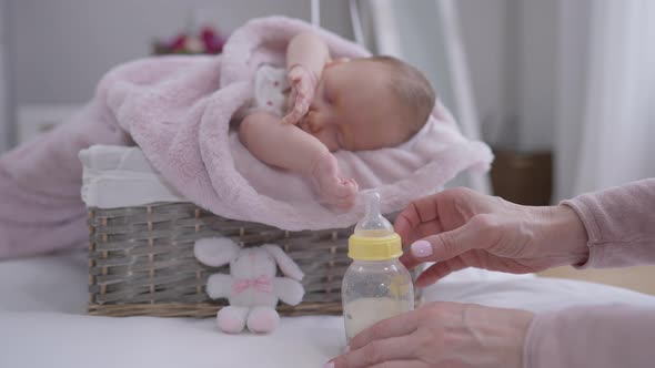 Female Hands Screwing Bottle Nipple with Cute Adorable Caucasian Infant Sleeping at Background